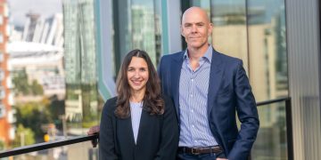 Picture of AbCellera’s Chief Technology Officer Dr. Veronique Lecault with CEO Dr. Carl Hansen at the company’s Mount Pleasant headquarters., outside on balcony. They are wearing dark blue blazers and light blue shirts.